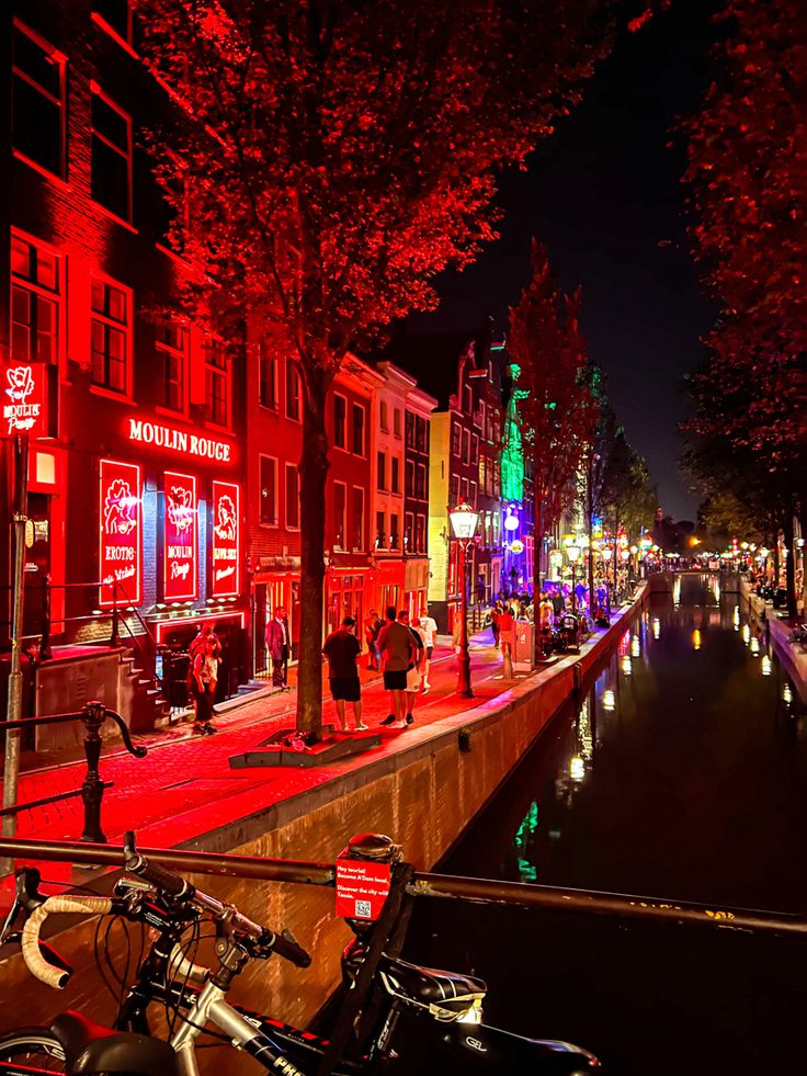 people walking down the street at night with red lights on buildings and bicycles parked in front of them