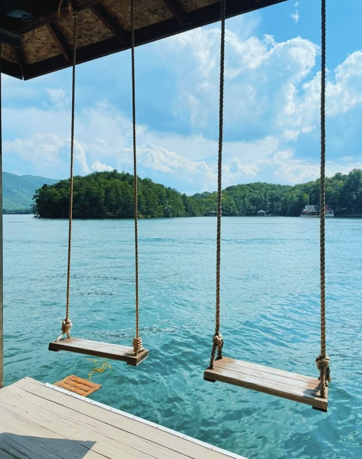 two wooden swings hanging from the side of a pier over water with trees in the background