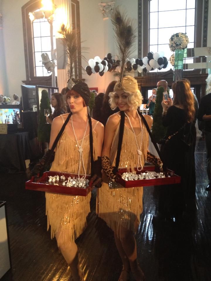 two women dressed in flappers and gats holding trays with food on them