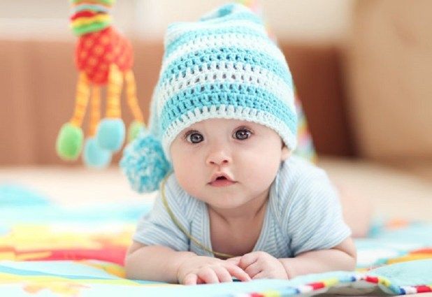 a baby laying on its stomach wearing a blue crocheted hat with pom poms