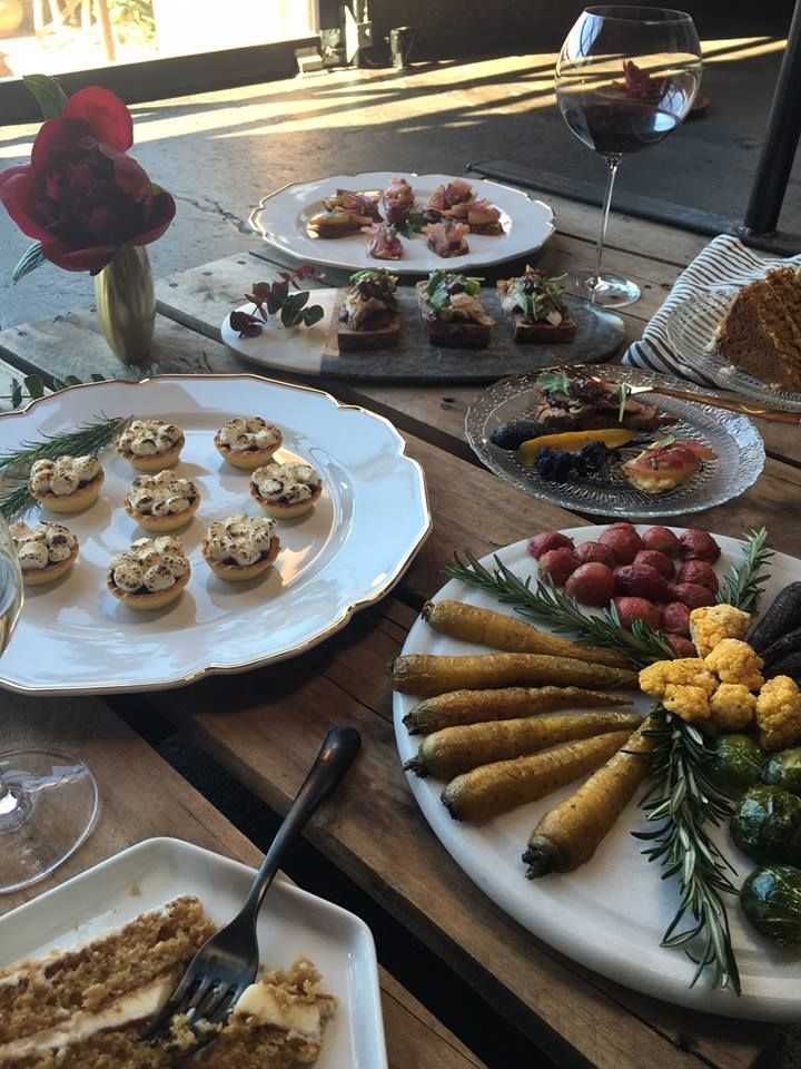 a table topped with plates and trays filled with food next to glasses of wine