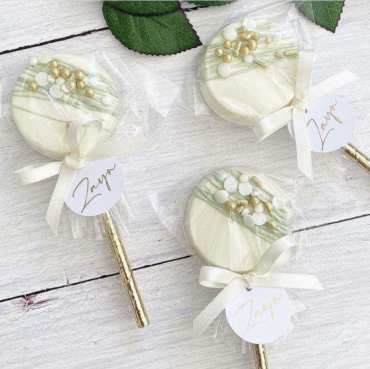 three white and gold candy lollipops with name tags on them sitting next to some green leaves