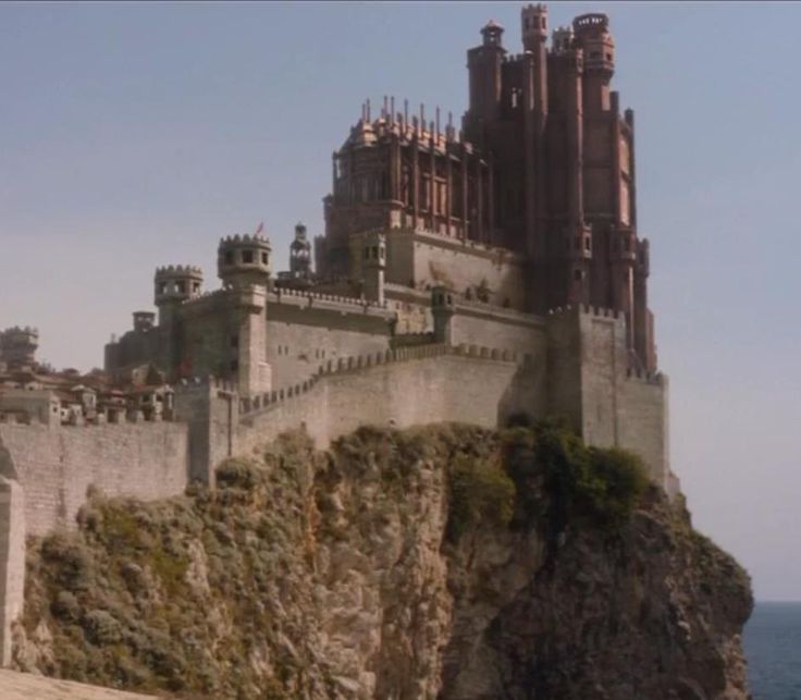 a man sitting on top of a cliff next to the ocean with a castle in the background