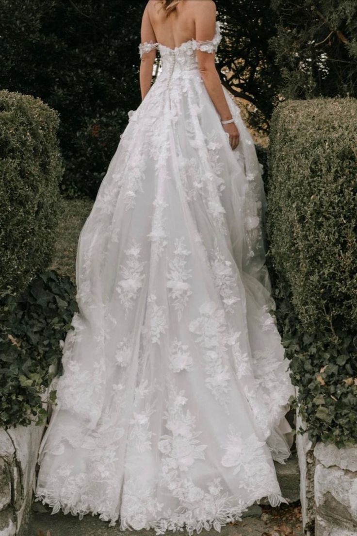 a woman in a white wedding dress standing next to some bushes and trees with her back turned towards the camera