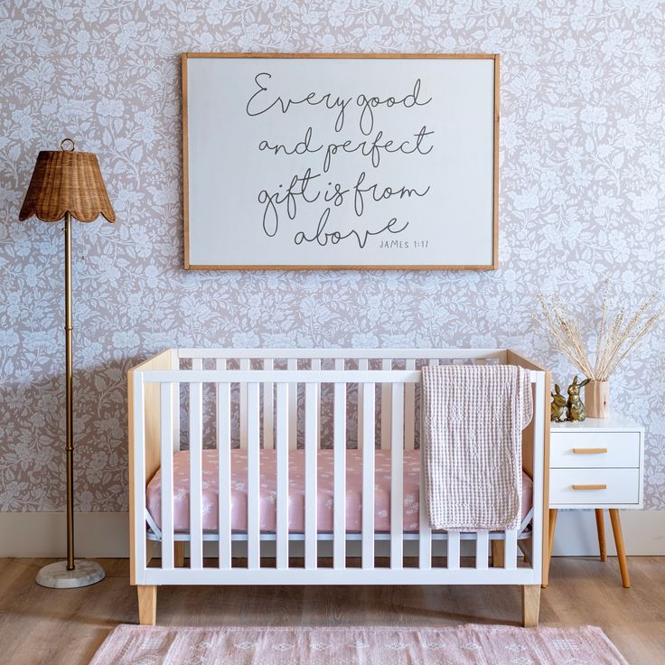 a white crib with pink bedding in front of a framed quote on the wall