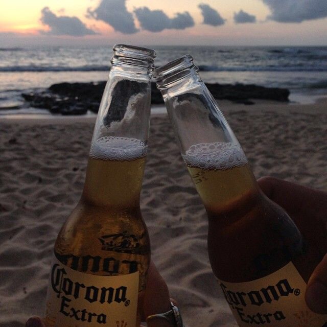 two bottles of corona beer are held up in front of the ocean and beach at sunset