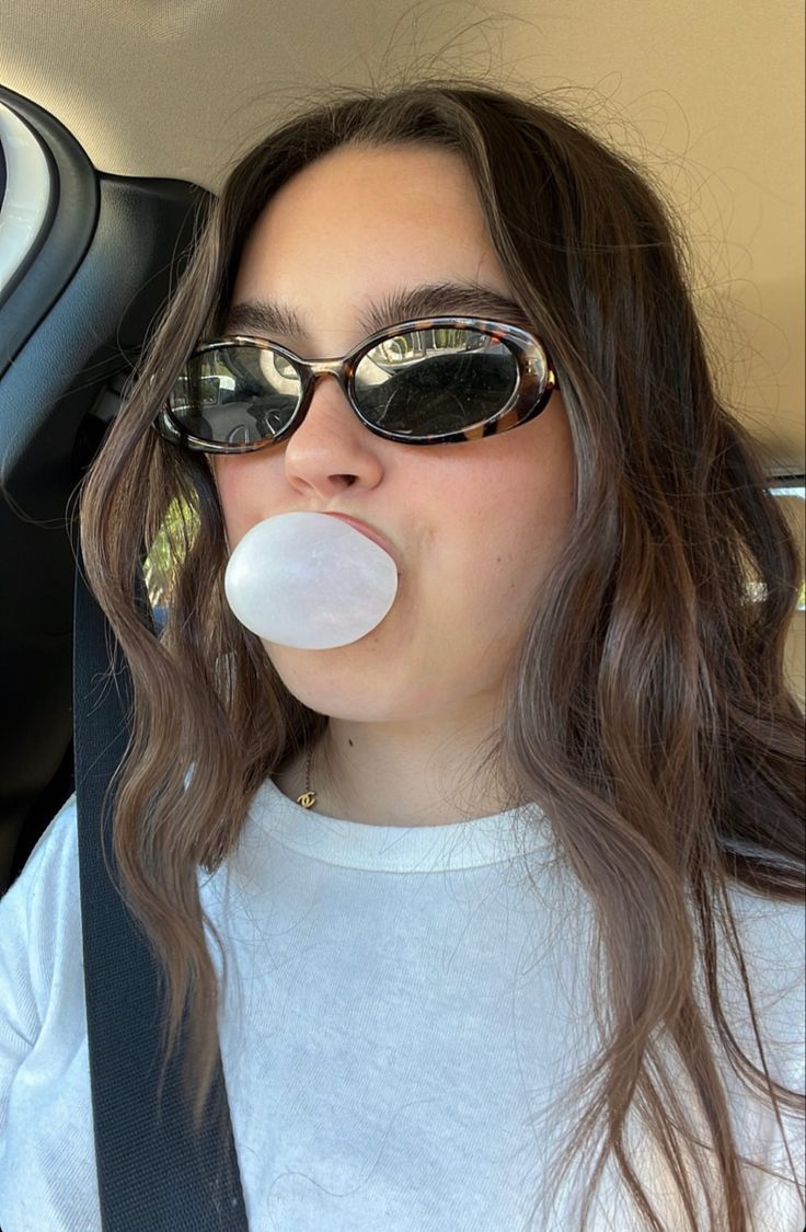 a woman with sunglasses blowing bubbles into her mouth in the back seat of a car