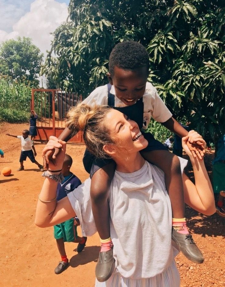 a woman carrying a child on her back in the middle of a dirt area with children playing around