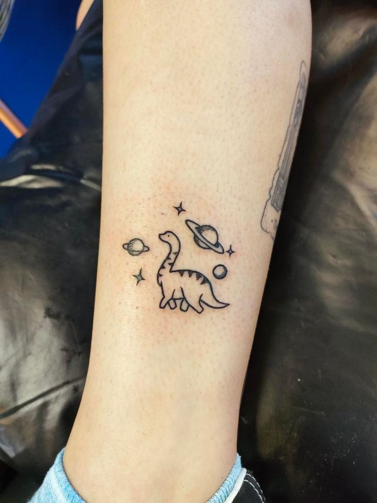 Small dinosaur tattoo in space. T Rex Space Tattoo, Cute Tattoos Dinosaur, Cute Dino Tattoo Matching, Dinasour Tattoo Matching, Cute Matching Dinosaur Tattoo, Star And Space Tattoos, Patchwork Dinosaur Tattoo, Cow On A Skateboard Tattoo, Dino Tattoos Cute