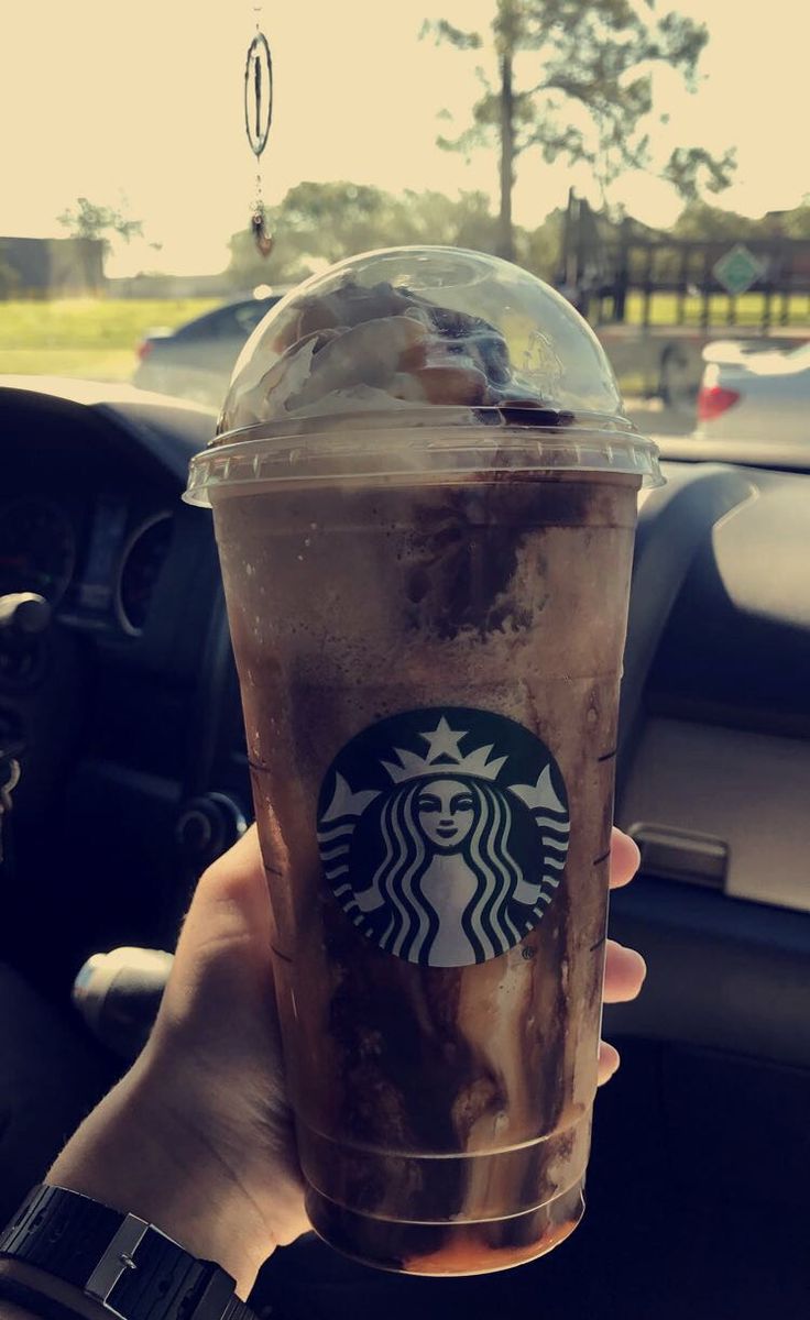 a person holding up a starbucks drink in their hand with the steering wheel behind them