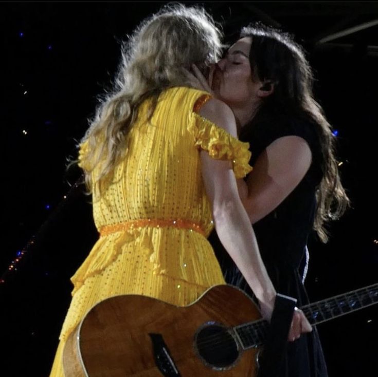 two women kissing each other while holding an acoustic guitar in front of them on stage