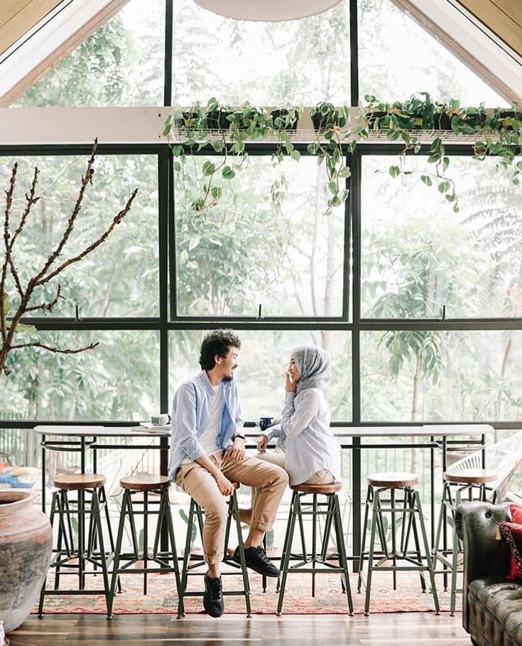 two people sitting at a table in front of a large window with plants on it