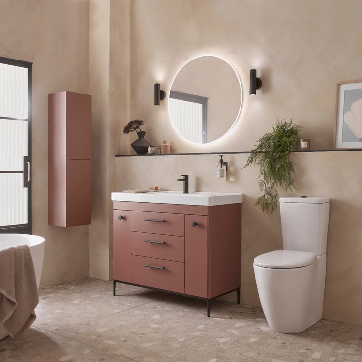 a white toilet sitting next to a sink in a bathroom under a round mirror on the wall