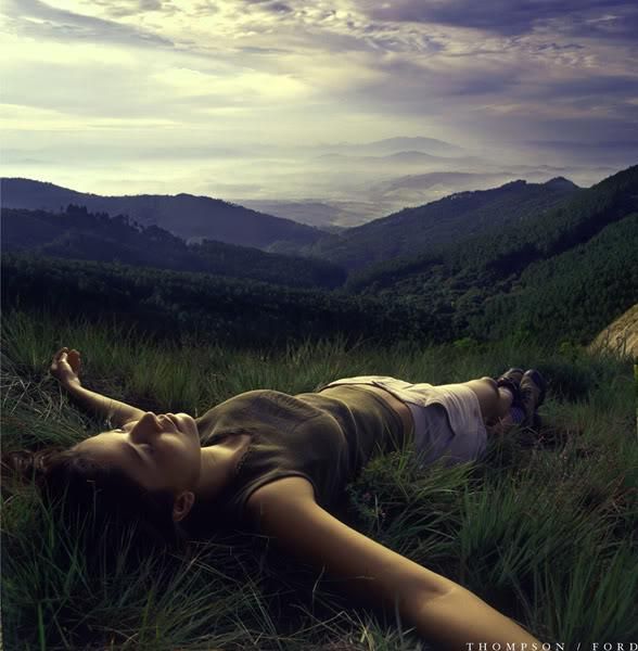 a woman laying down in the grass with her eyes closed and mountains in the background