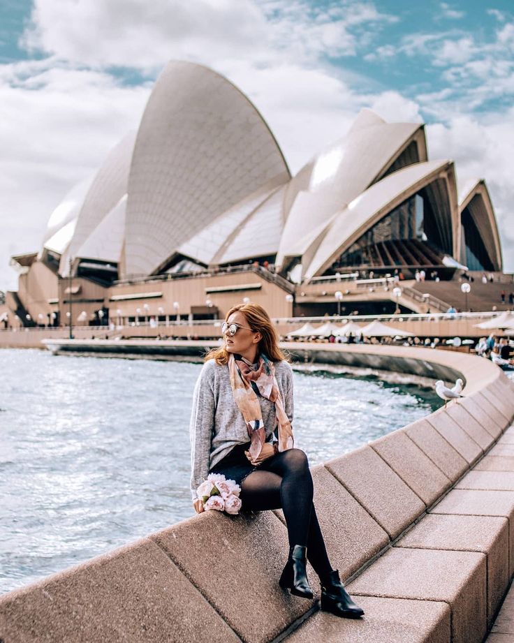 a woman sitting on the edge of a wall next to water and a large building