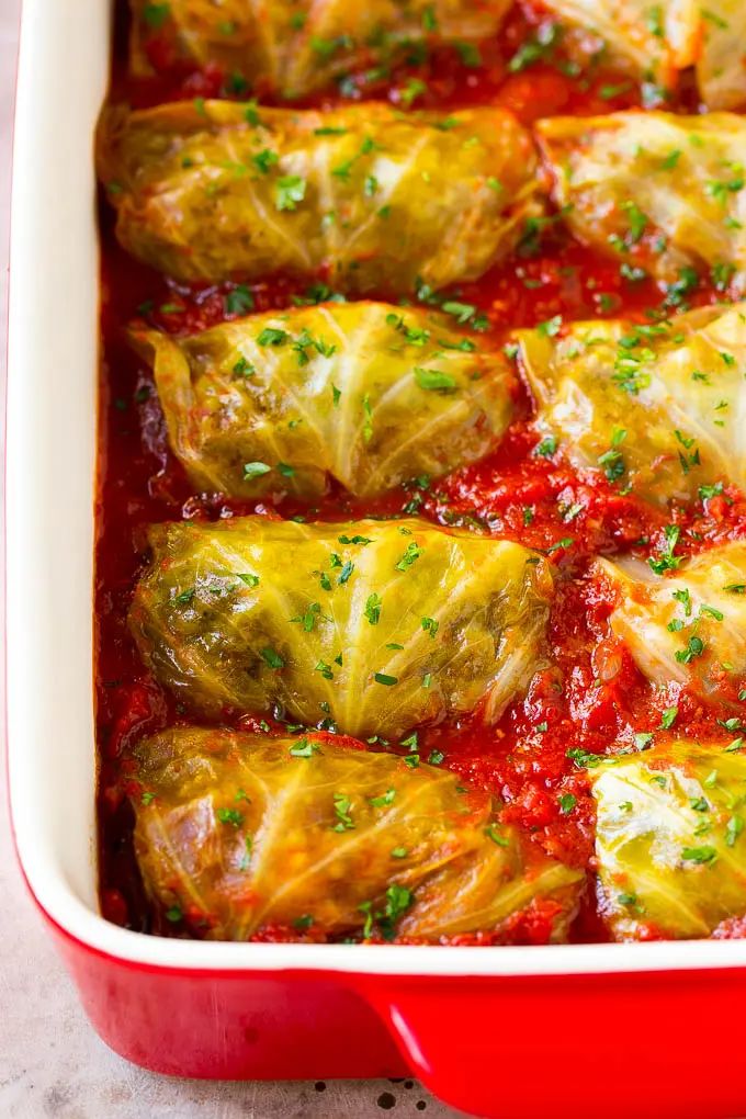 a red casserole dish filled with stuffed cabbage rolls and marinara sauce, garnished with parsley