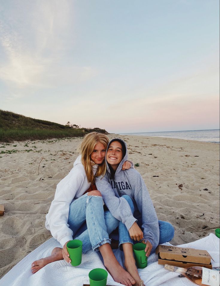 two women are sitting on the beach with cups in their hands and one is hugging her