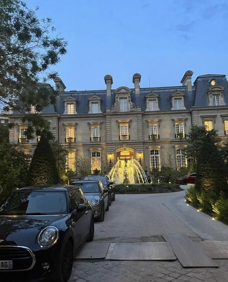 cars are parked in front of a large building at night with lights on the windows