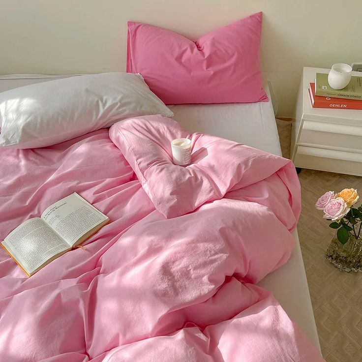 a bed with pink sheets, pillows and a book on top of it next to a vase filled with flowers