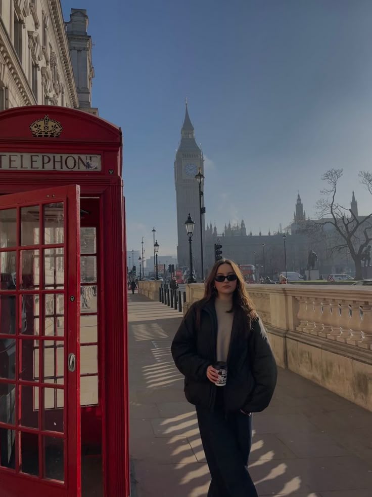 a woman standing next to a red phone booth on the side of a road with a clock tower in the background