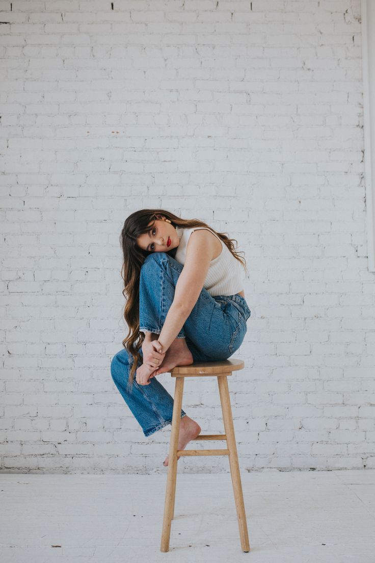 a woman sitting on top of a wooden stool in front of a brick wall with her legs crossed