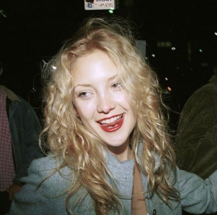 a woman with blonde hair and red lipstick smiling at the camera while standing in front of a group of people