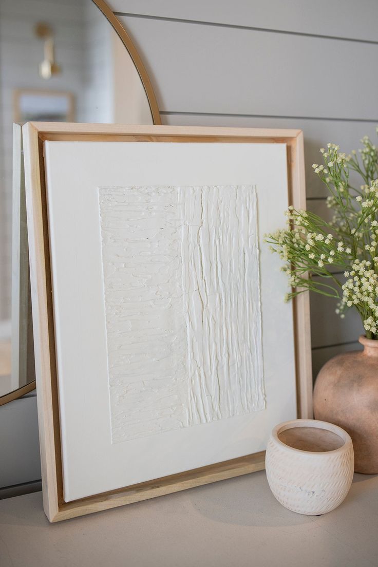 there is a vase with flowers next to a framed art piece on the table in front of a mirror