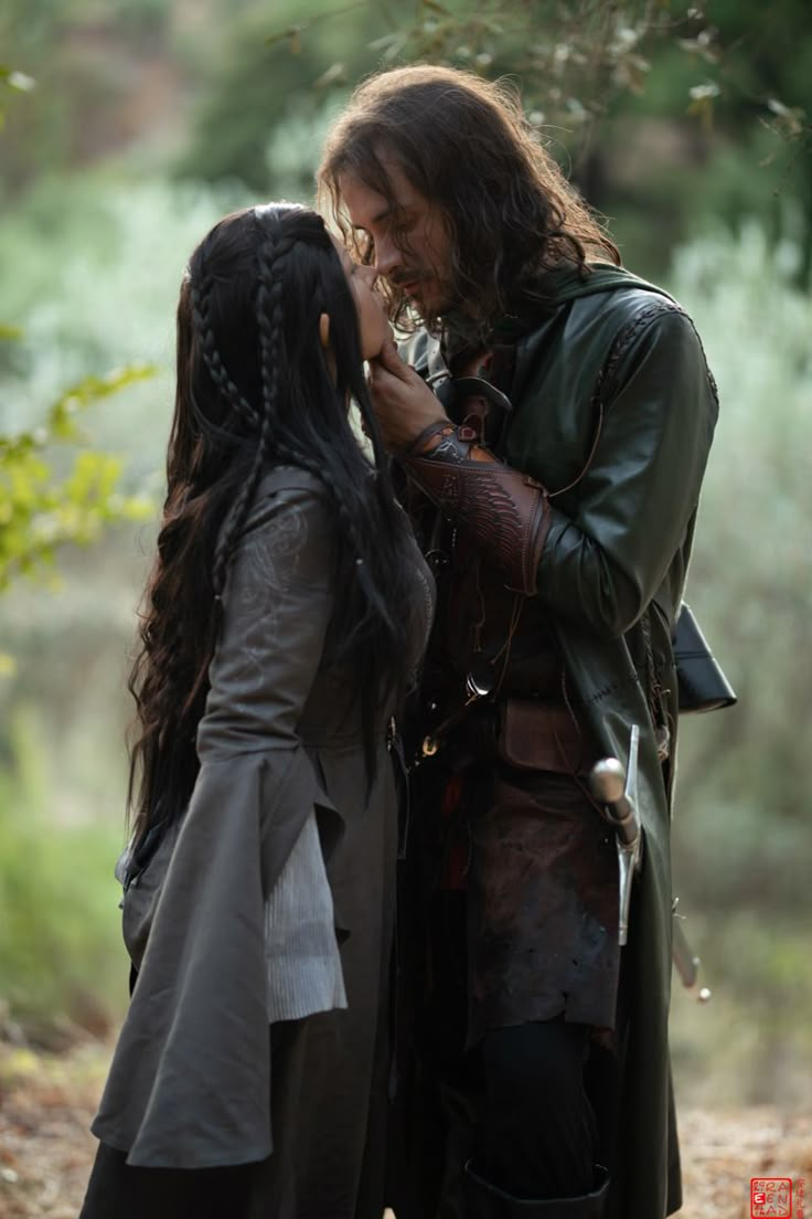 two people dressed in medieval clothing standing next to each other on a dirt road with trees in the background