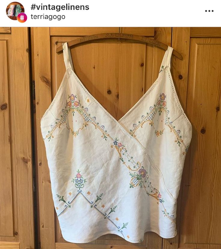 a white top with embroidered flowers on it hanging from a wooden hanger in front of wood paneled doors