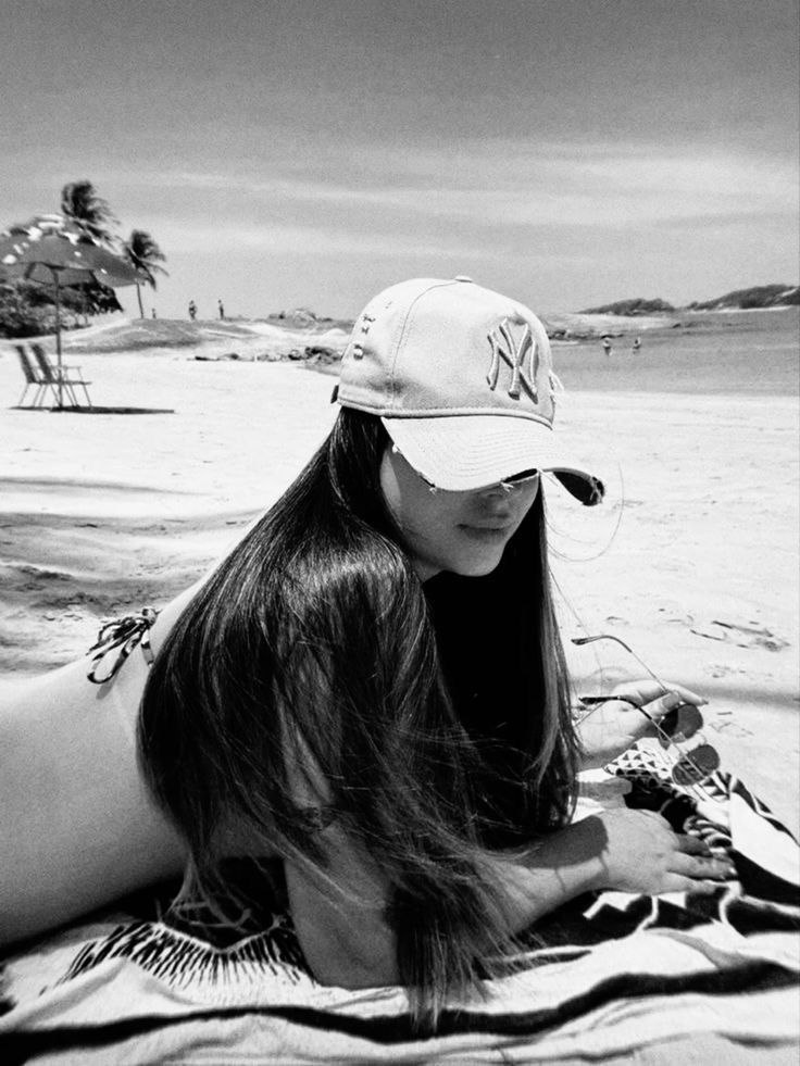 a woman laying on top of a beach next to the ocean wearing a baseball cap