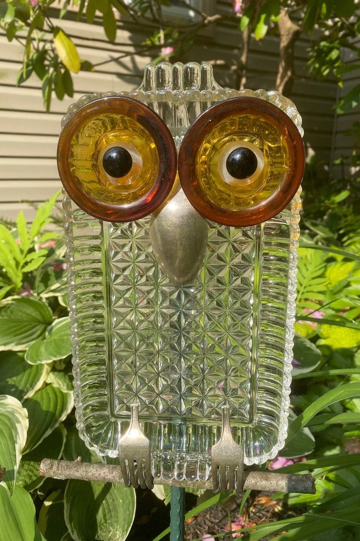 an owl made out of glass sitting on top of a metal stand in front of some plants