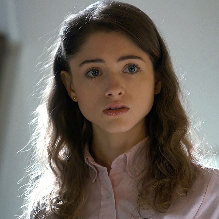 a woman with long brown hair wearing a white shirt and looking at the camera while standing in front of a mirror