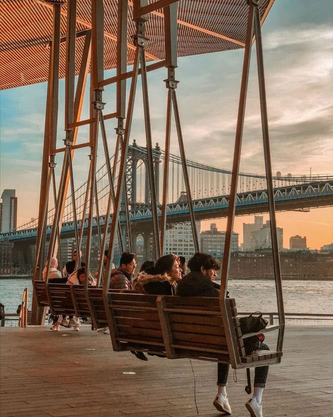 people are sitting on swings in front of the water and a bridge with a blue sky behind them