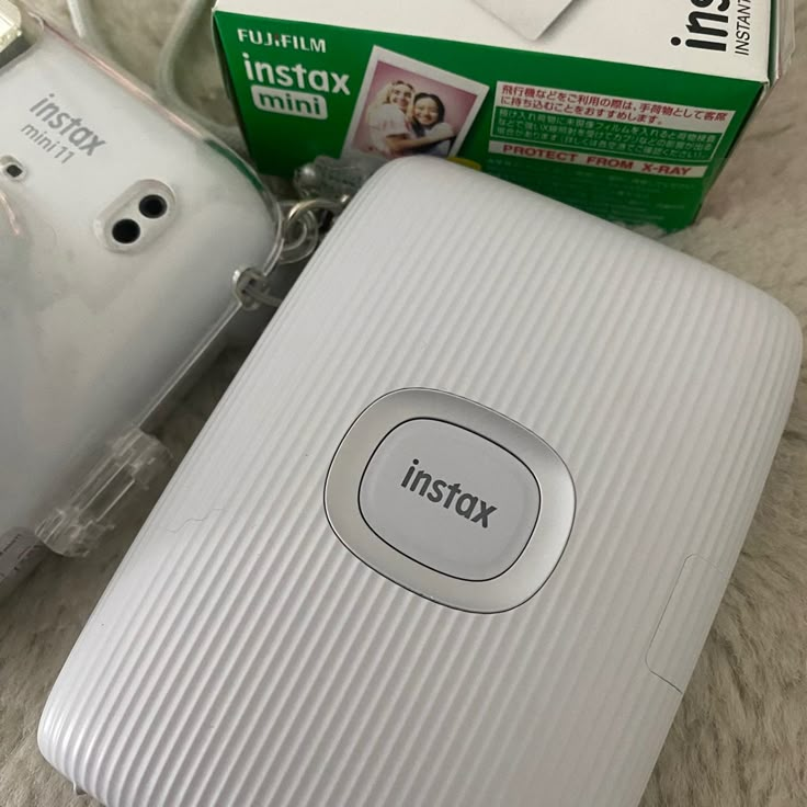 a white camera sitting on top of a bed next to a box with the word instax printed on it