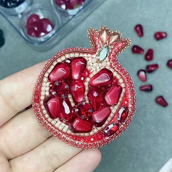 a hand holding a red pomegranate shaped brooch with jewels on it