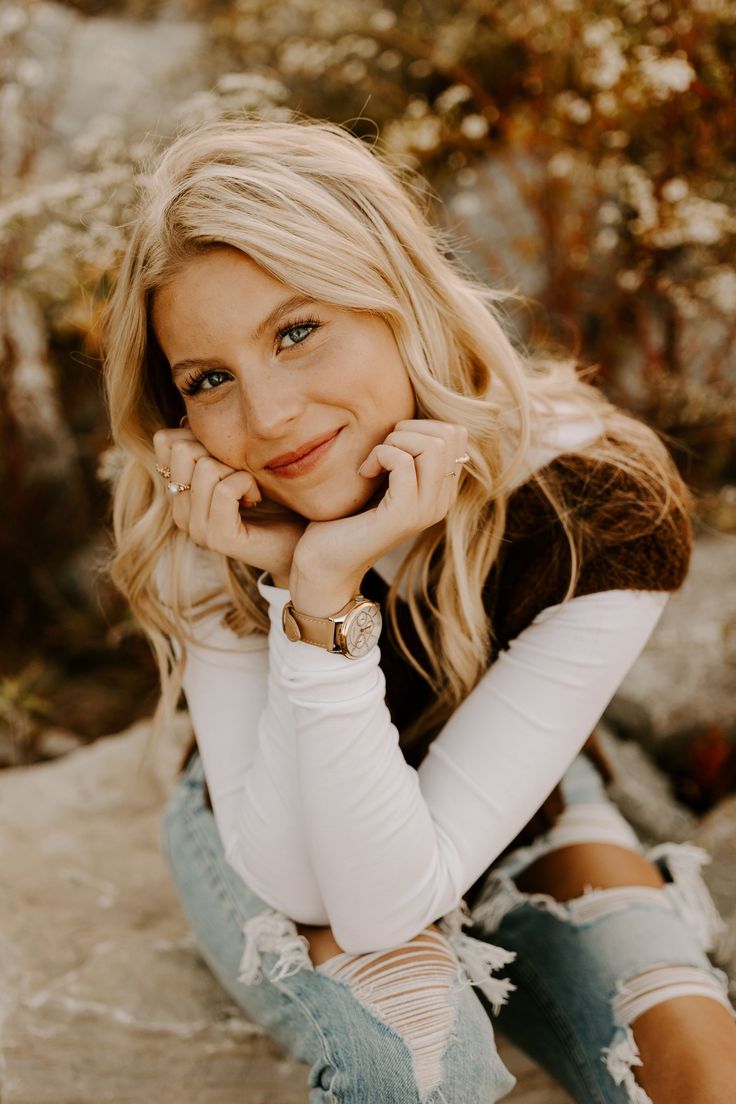 a blonde woman sitting on top of a rock next to trees and bushes with her hand under her chin