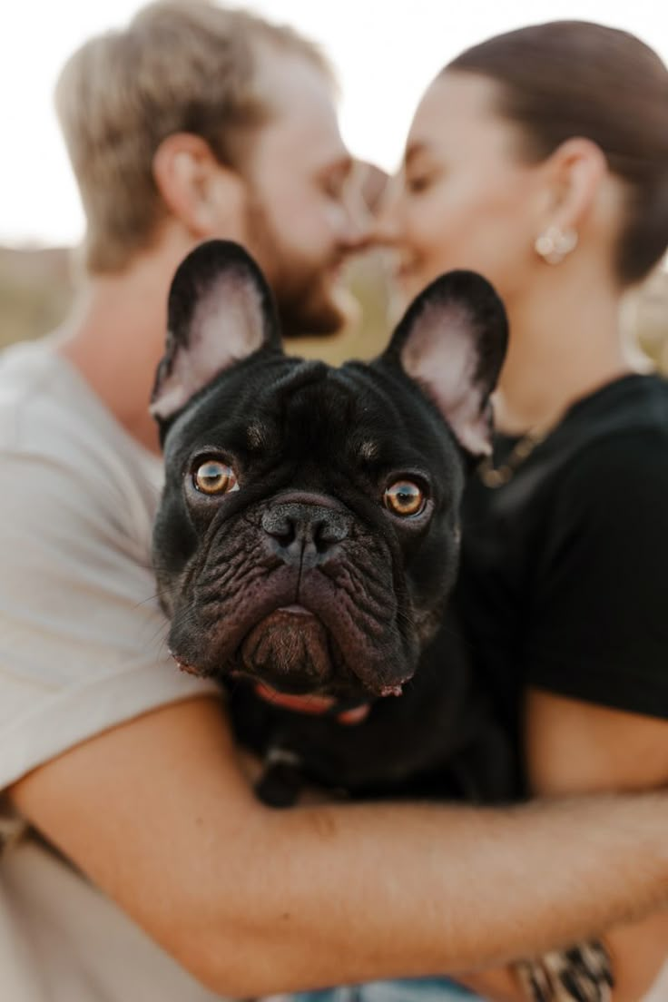 a man and woman kissing while holding a black dog in their arms, with the dog looking at the camera