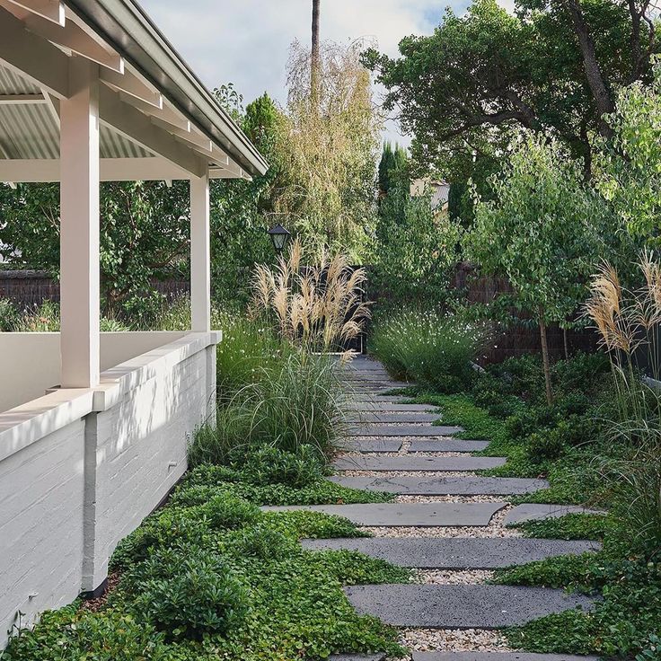 a stone path in front of a white house surrounded by trees and bushes with grass growing on both sides