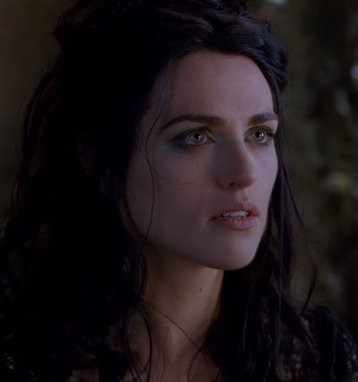 a woman with dark hair and blue eyes stares into the distance while standing in front of a wall