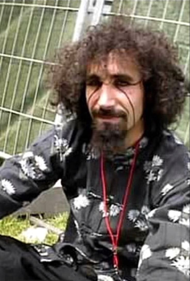 a man with long hair sitting in front of a metal fence and looking at the camera