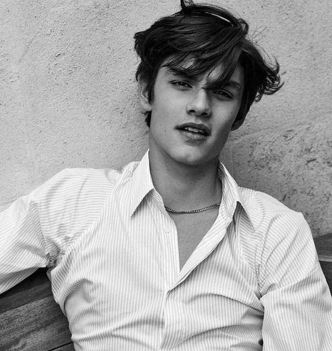 a young man sitting on top of a wooden bench next to a wall and wearing a collared shirt