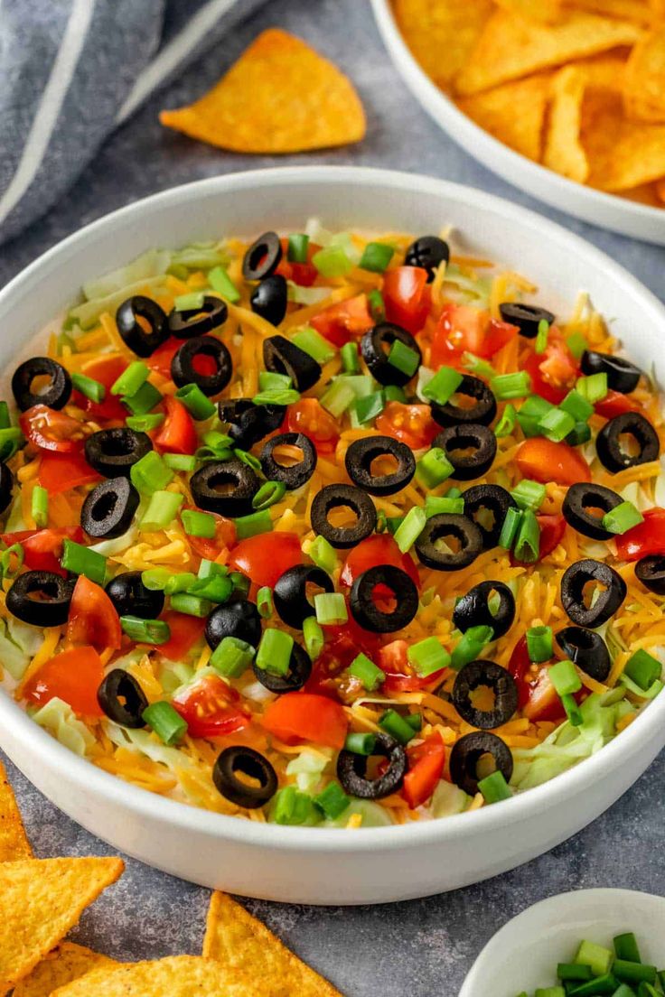 a white bowl filled with black olives, peppers and cheese surrounded by tortilla chips