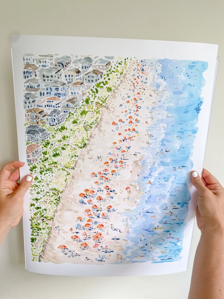 two hands holding up a watercolor drawing of a beach with houses in the background