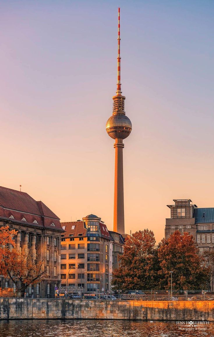 the television tower in berlin, germany is lit up at sunset as seen from across the river