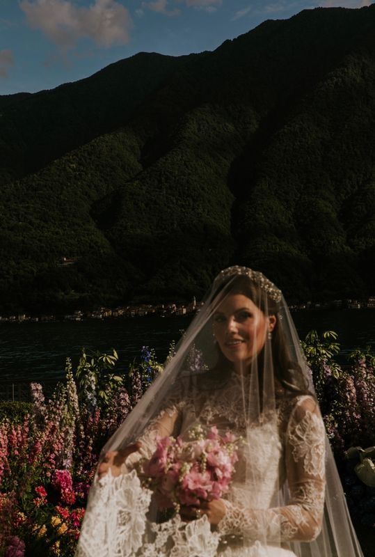 a woman in a wedding dress and veil standing next to flowers with mountains in the background