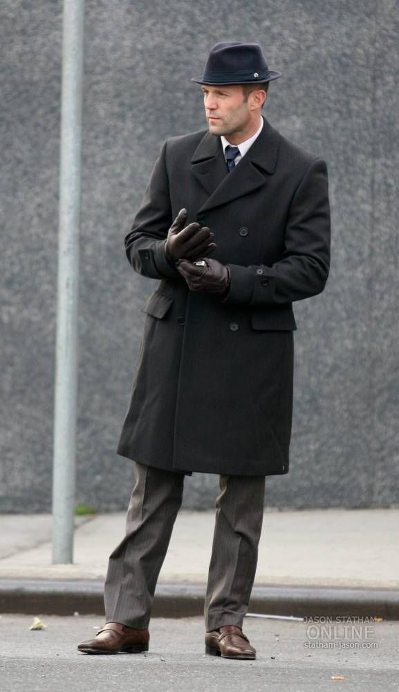 a man in a black coat and hat standing on the street with his hands folded