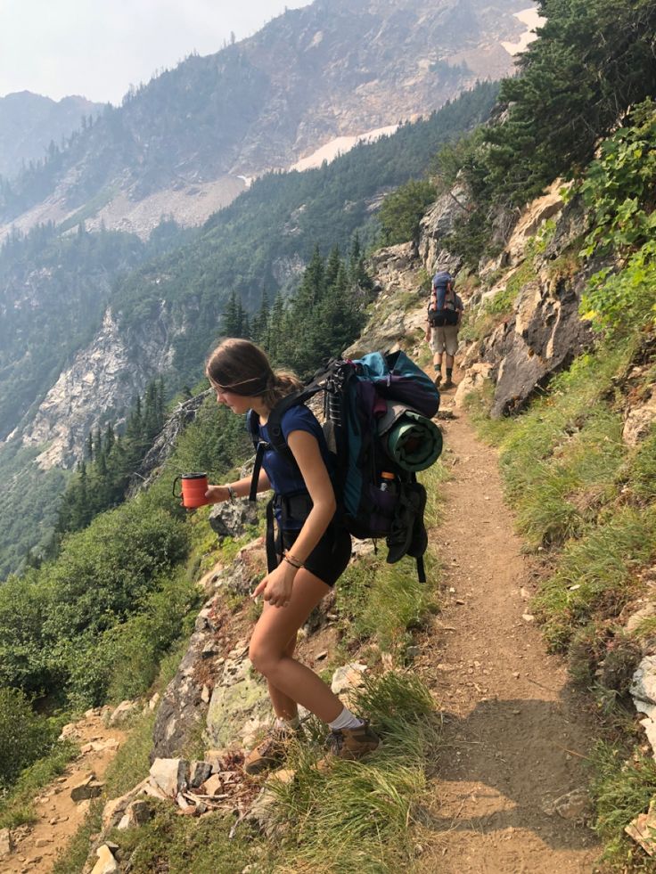 a woman hiking up the side of a mountain with a backpack and cup in her hand