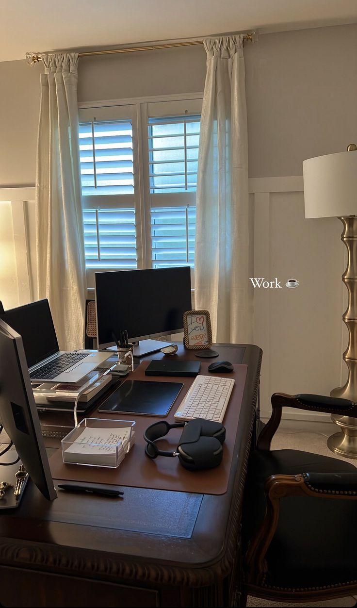 a desk with two computers on it in front of a window that has blinds and drapes