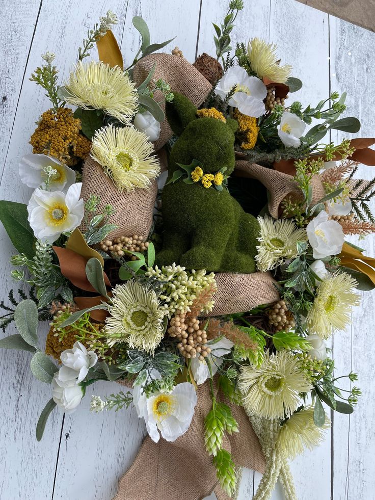 a teddy bear wreath with flowers and greenery on a white wooden surface, ready for someone to put them in