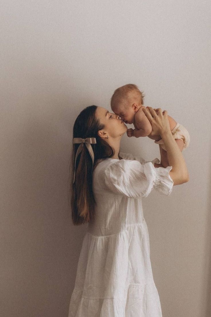 a woman holding a baby up to her face and kissing it's cheek while standing in front of a white wall